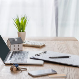 wooden desk with laptop, smartphone, house keys, clipboard with pen, house model and green plant